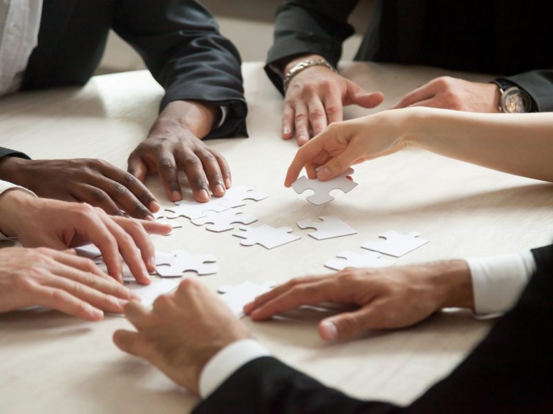 Close up of multiethnic team solving blank puzzle on table. Female hand offering puzzle piece, problem solution concept. Multi-racial executive employees brainstorming about work related problem.